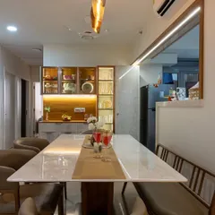 The dining area is strategically placed near the open kitchen, featuring a beautiful marble-top table and a custom-lit crockery unit that adds a touch of elegance and provides ample storage.