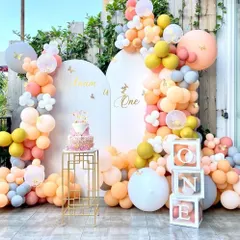 An elegant pastel-themed first birthday party for "Anaan." This outdoor setup features two arched backdrops, a beautiful organic balloon garland, a modern cake stand, and "ONE" letter blocks.