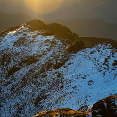 A drone photograph capturing the golden light of the sun hitting a snow-dusted peak in Uttarakhand. This image highlights the textures of the rock and snow, showing the raw beauty of the mountains during the golden hour.