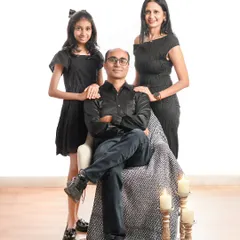 An elegant and classic family portrait. The father is seated while his wife and daughter stand beside him, all dressed in sophisticated black attire in a warmly lit studio.