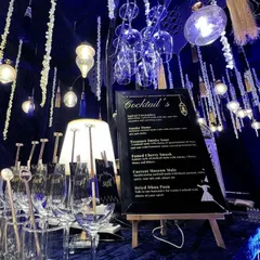 A detailed shot of the Sufi bar, showcasing the custom cocktail menu, personalized 50th-anniversary stirrers, and the warm glow from hanging Edison bulbs and a table lamp.
