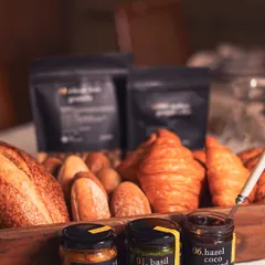 A selection of our artisanal pantry products, including seasonal jam, basil pesto, and hazel coco spread, alongside fresh breads and our wheat-free granola. These are the building blocks of a gourmet breakfast.