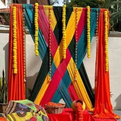 A creative and modern backdrop design using a criss-cross pattern of colorful drapes in orange, yellow, teal, and black. This geometric approach to fabric draping is accented with marigold strings for a fusion look.
