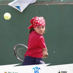 A young girl in a red bandana unleashes a powerful backhand during a match. Her determined expression shows the competitive spirit we love to see on our courts.