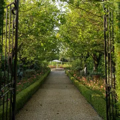 An inviting entrance through a wrought iron gate into a formal garden. The gravel path is flanked by low, clipped boxwood hedges and colorful flower borders, leading to a focal point in the distance.