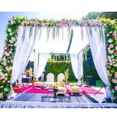 A beautiful outdoor mandap setup with a full floral arch and white drapery. The stage is set on a vibrant pink carpet, adding a pop of color to the otherwise classic and elegant design.