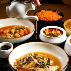 An action shot capturing a delicate drizzle over a bowl of Asian soup. The composition, with a spread of other dishes, creates a dynamic and appetizing scene that highlights the textures and flavors of the cuisine.