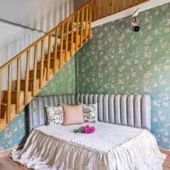 A whimsical bedroom nook designed under a wooden staircase. The custom-made daybed with a channel-tufted headboard is set against a lovely floral wallpaper, creating a magical little retreat.