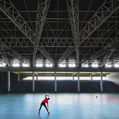 Inside the "Figured Ground" sports arena, a lone athlete stretches on the court. The vast, column-free space is made possible by the complex steel truss roof structure, which spans the entire arena.