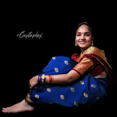 This full-length seated pose showcases the complete traditional Marathi ensemble, from the elegant drape of the blue silk saree to the delicate anklet. The dark background ensures the vibrant attire is the main focus.
