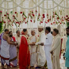 A beautiful moment during a traditional South Indian wedding ceremony, framed by the elegant mandap. The decor features hanging garlands of white tuberose, accented with pink lotus buds and red roses.