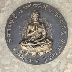 This circular relief sculpture features Lord Buddha in the 'Abhaya Mudra' pose, surrounded by the 'Om Mani Padme Hum' mantra. The piece has a dual-tone bronze and grey finish.