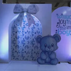 A close-up of a baby boy shower backdrop. This setup features a large gift box design with a floral pattern, a cute teddy bear cutout, and the phrase "so much joy in this little boy".