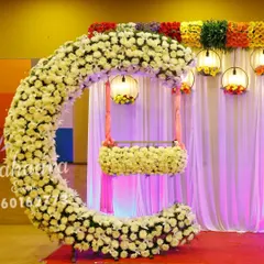 A stunning cradle ceremony setup featuring a crescent-shaped arch made of white flowers. The hanging cradle, also adorned with flowers, creates a beautiful focal point, while colorful lanterns add a festive touch.