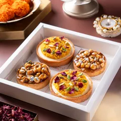 A close-up of four exquisite mini tarts in a white box, two topped with motichoor and pistachio, and two with a nutty caramel glaze. These fusion desserts are a perfect example of how I blend traditional Indian flavors with classic pastry techniques.