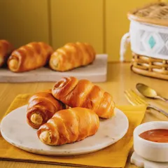 Golden-baked sausage rolls arranged on a marble plate. The soft lighting and warm yellow background create a cozy and inviting scene, perfect for a bakery's breakfast menu.
