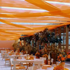An outdoor dining setup for a Mamera ceremony, with tables adorned with orange floral centerpieces under a canopy of flowing orange drapes.