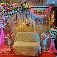A festive Mehndi stage setup combining a lush floral arch with bright pink drapes and fairy lights. The 'Mehndi' sign in Hindi adds a traditional touch to this vibrant and colorful decoration.