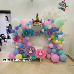 A magical unicorn-themed party backdrop, perfect for a child's birthday or a baby welcome. This setup includes a multi-colored pastel balloon arch, a large unicorn foil balloon, and charming paper fan details.