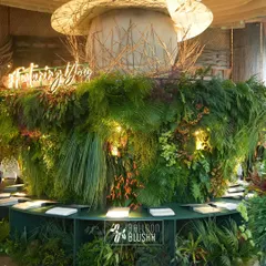 A massive, circular bar installation for home decor brand Nesterra, completely enveloped in lush, dense greenery. The brand's illuminated logo and hashtag serve as a focal point, creating an unforgettable brand activation and photo opportunity for guests.