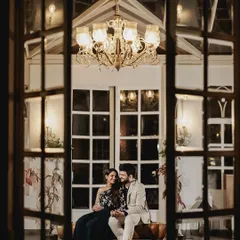 An evening shot through a window, capturing a couple sharing a quiet moment inside a beautifully lit cafe. This perspective creates a feeling of watching a private, romantic scene unfold.