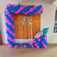 A baby shower setup for a home party. I created a photo corner with a gold tinsel curtain, a pink and blue balloon frame, and a 'Baby Shower' banner.
