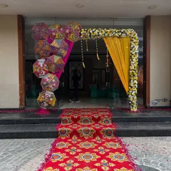 A beautiful entrance for a banquet hall, featuring a floral arch with yellow drapes, Rajasthani umbrellas, and a traditional patterned carpet.
