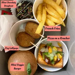 A close-up of a perfectly portioned snack box for a kid's party. It includes a mini veggie burger, veg lollipops, french fries, pizza on a cracker, and a mini chocolate doughnut, a complete and happy meal for any child.