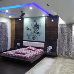 A dramatic bedroom design featuring a floating bed and a large, back-painted glass panel with a floral etching behind the headboard. The false ceiling incorporates blue cove lighting, adding a cool, ambient glow to the room.