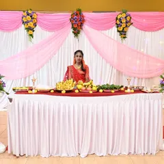 An elegant stage for a half-saree ceremony. The backdrop uses simple white and pink drapes, accented with colorful floral bouquets on pillars, creating a graceful setting.