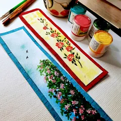 A pair of hand-painted bookmarks, perfect for any book lover. One features a delicate rose vine, and the other a lush bougainvillea against a blue sky, both painted with acrylics on watercolor paper.
