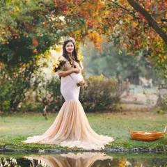 An expectant mother in a lovely mermaid-style gown stands by a pond in a park with autumn foliage. The warm colors of the background beautifully complement her gentle and radiant look.