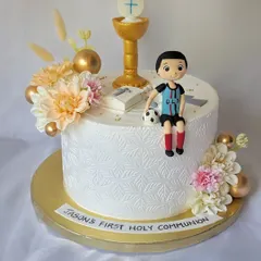 A First Holy Communion cake for Jason. The elegant white design is stenciled and decorated with a chalice, a Bible, and a figurine of the boy in his football jersey.
