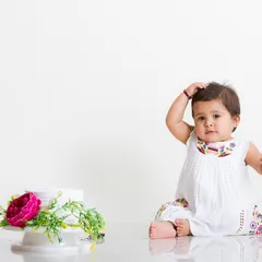 Simple, clean, and classic. A baby in a lovely white dress sits next to a small floral arrangement, looking thoughtfully at the camera. This minimalist style puts all the focus on your child's sweet personality.