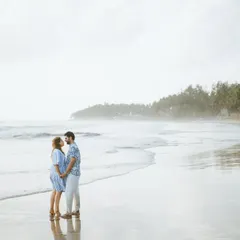 A wide-angle shot of the couple standing on a wet beach, with the waves receding around them. The misty, atmospheric background and their quiet stance create a serene and romantic scene.