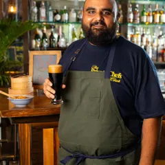 A proud portrait of Chef Harsh at Common House Karjat, holding a glass of craft beer. This shot connects the chef directly to the product, presenting him as a welcoming host and the expert behind the pop-up menu.