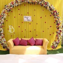 A simple yet elegant mehndi backdrop featuring a large floral ring against a bright yellow curtain. The sofa provides comfortable seating, and the setup can be personalized with the couple's names.