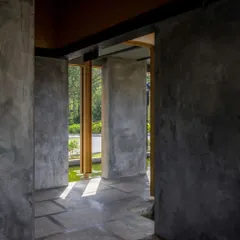 The interior of the Teyla project, where raw micro-concrete walls and stone floors create a cool, cave-like passage. Sunlight filters in, highlighting the textures and creating a path that leads the eye towards the green pond outside.