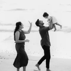 A candid black and white shot of a family playing on the beach. The father lifts the baby high, and the mother looks on with a smile, capturing a moment of pure, unfiltered happiness.