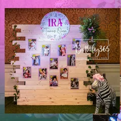 A personalized photo memory wall from a jungle-themed first birthday. Photos from Ira's first year are displayed on a rustic wooden backdrop, accented with climbing vines and a custom light-up sign.