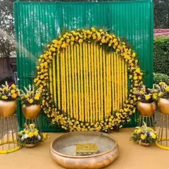 A classic Haldi ceremony ring backdrop for an outdoor setting. The design features a full circle of marigolds with hanging flower strings, set against a green curtain, with a traditional urli in front.