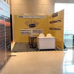 An exhibition booth for Alpha Omega Enterprises, designed to showcase product catalogs and information clearly. The layout includes a small, semi-private meeting desk for discussions with potential clients.