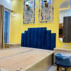 A bold and beautiful master bedroom design. The royal blue velvet headboard stands out against the vibrant yellow wall, which is decorated with framed panels and classic wall lights.