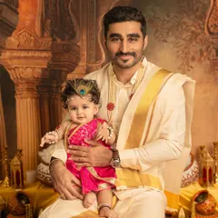 A proud father holds his baby, who is dressed as little Krishna for a festive photoshoot. The traditional outfits and detailed set design make this a special portrait.