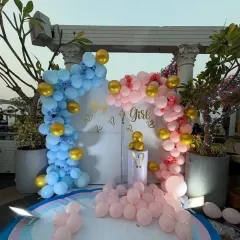 An elegant outdoor gender reveal party on a terrace. The "Boy or Girl?" backdrop is framed by two beautiful balloon arches in pastel pink and blue with gold accents.