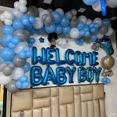 A close-up of a "Welcome Baby Boy" wall decoration. The setup features blue foil letter balloons surrounded by an arch of blue, grey, and white balloons, perfect for a bedroom or living room wall.
