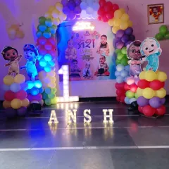 This is a closer look at a Cocomelon first birthday party setup. It features a custom backdrop with the baby's photos, character cutouts of JJ and his friends, and marquee lights spelling out the birthday boy's name.