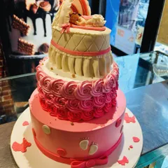 A three-tier pink baby shower cake with a quilted bassinet topper holding a sleeping baby.