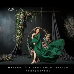 An elegant studio portrait featuring a woman in a flowing green gown on a floral-adorned swing. The dark, moody background creates a sophisticated and artistic atmosphere.