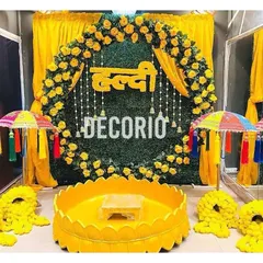 A bright and sunny Haldi setup with a circular green backdrop adorned with yellow roses and a 'Haldi' cutout. The decor is completed with traditional colorful umbrellas and marigold arrangements, creating a joyful ambiance.